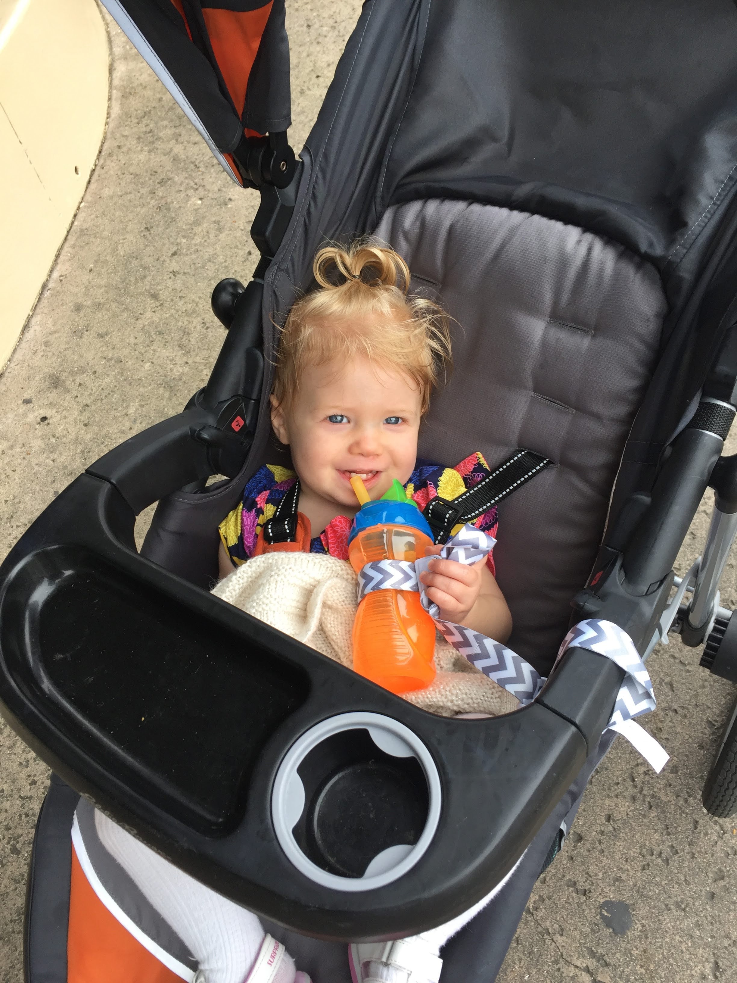 An infant hold a sippy cup in a stroller; the cup is attached to the stroller with a tether.