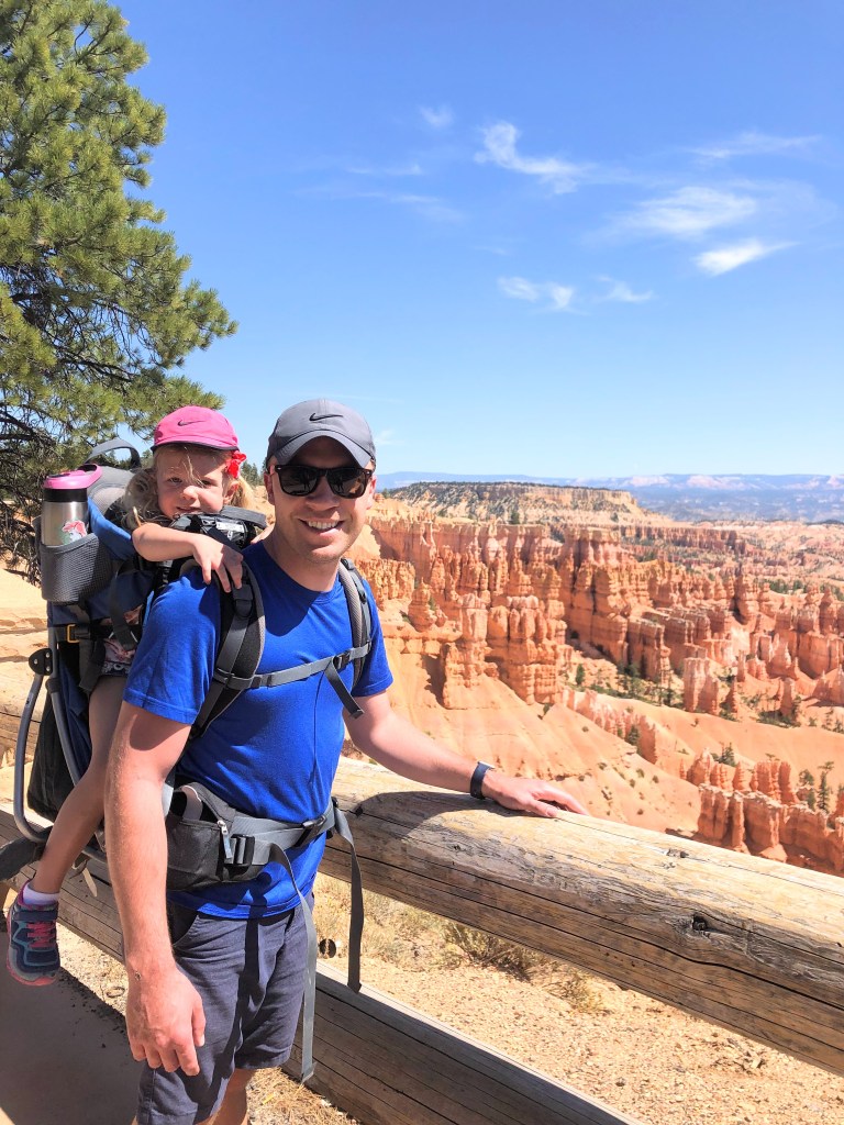 Man with child in hiking backpack on his bike, both smiling at the camera next to Bryce Canyon
