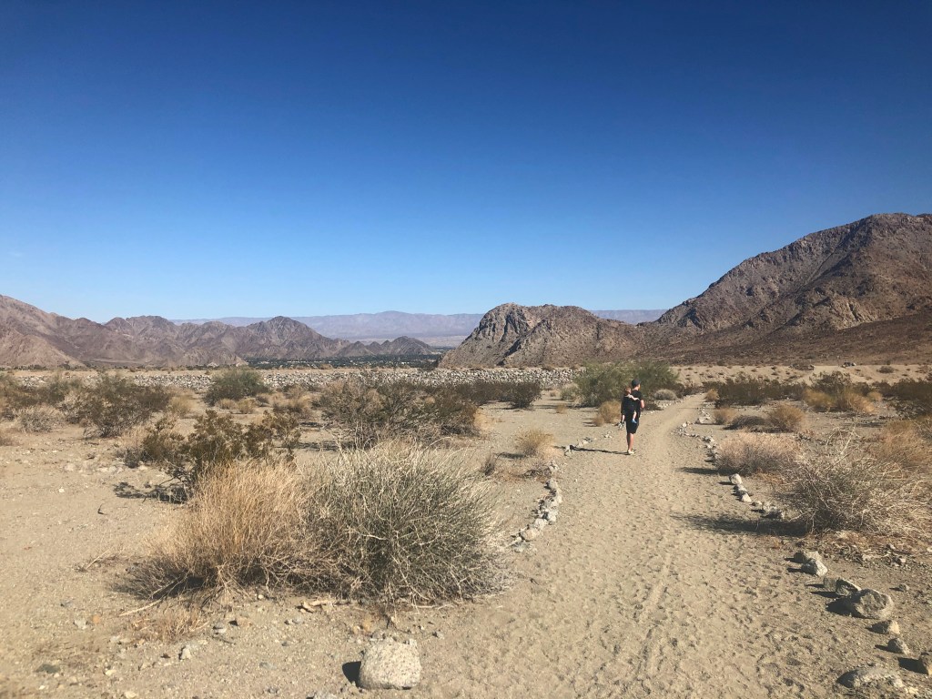 Desert trail surrounded by mountains with a man carrying a baby on his back